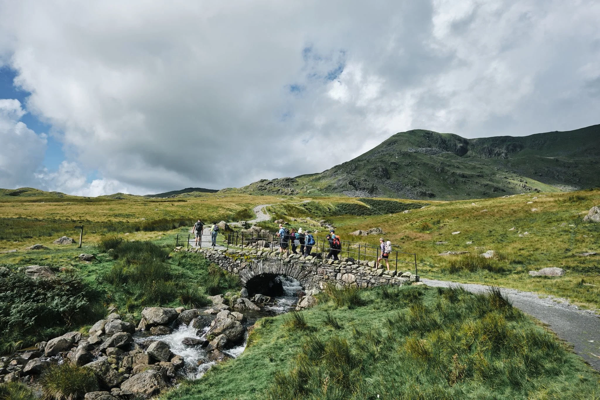 A group of hikers crossing a small stone bridge over a stream in a lush green mountain landscape with cloudy sky.