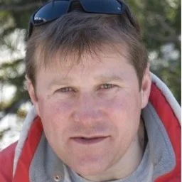 Close-up of a man outdoors, wearing sunglasses on his head and a gray jacket with red accents.
