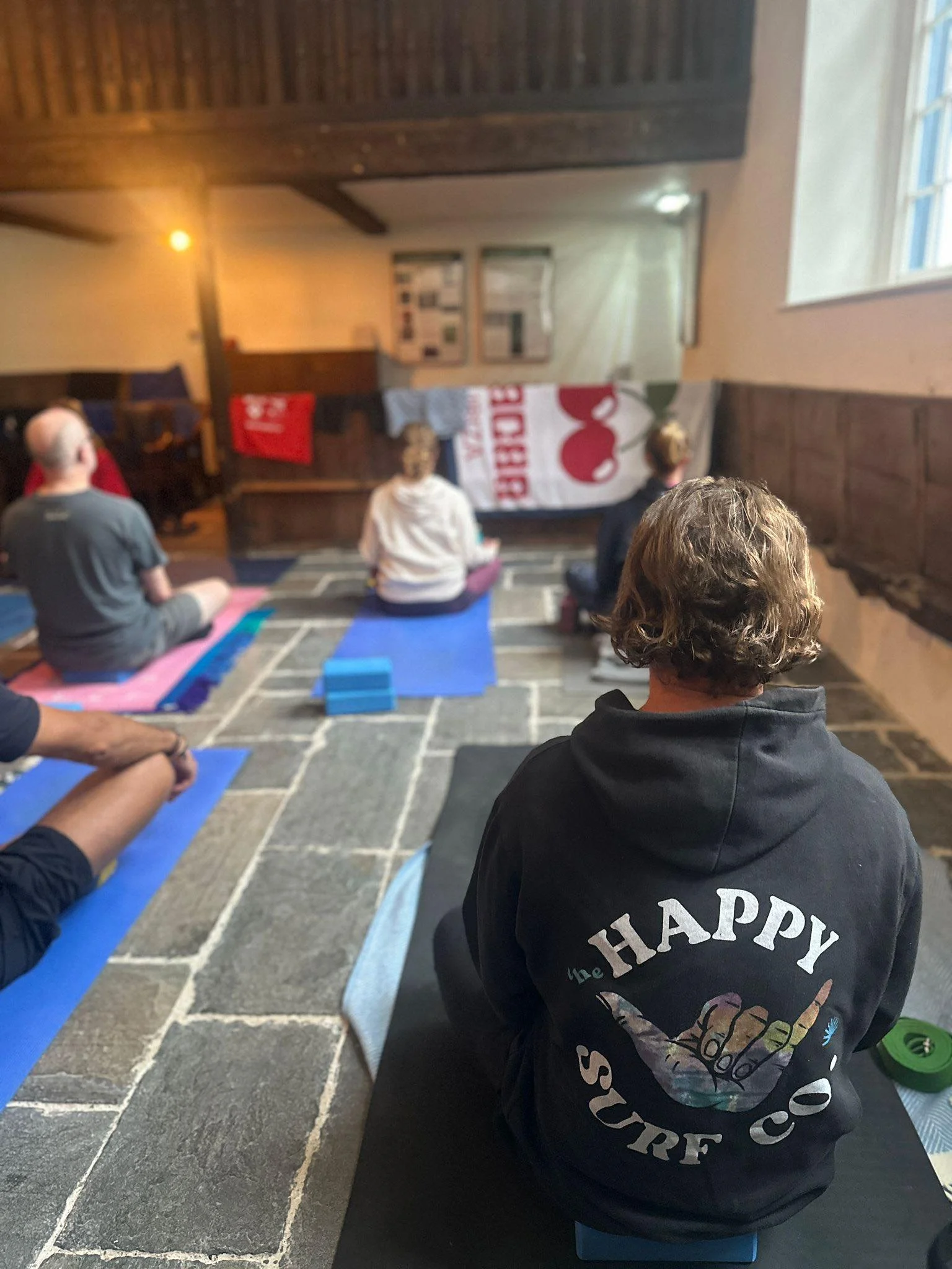 People participating in a yoga or meditation class in a cozy, rustic room with stone floor, wooden beams, and natural light coming through a window. Some individuals are sitting on mats in a cross-legged position.