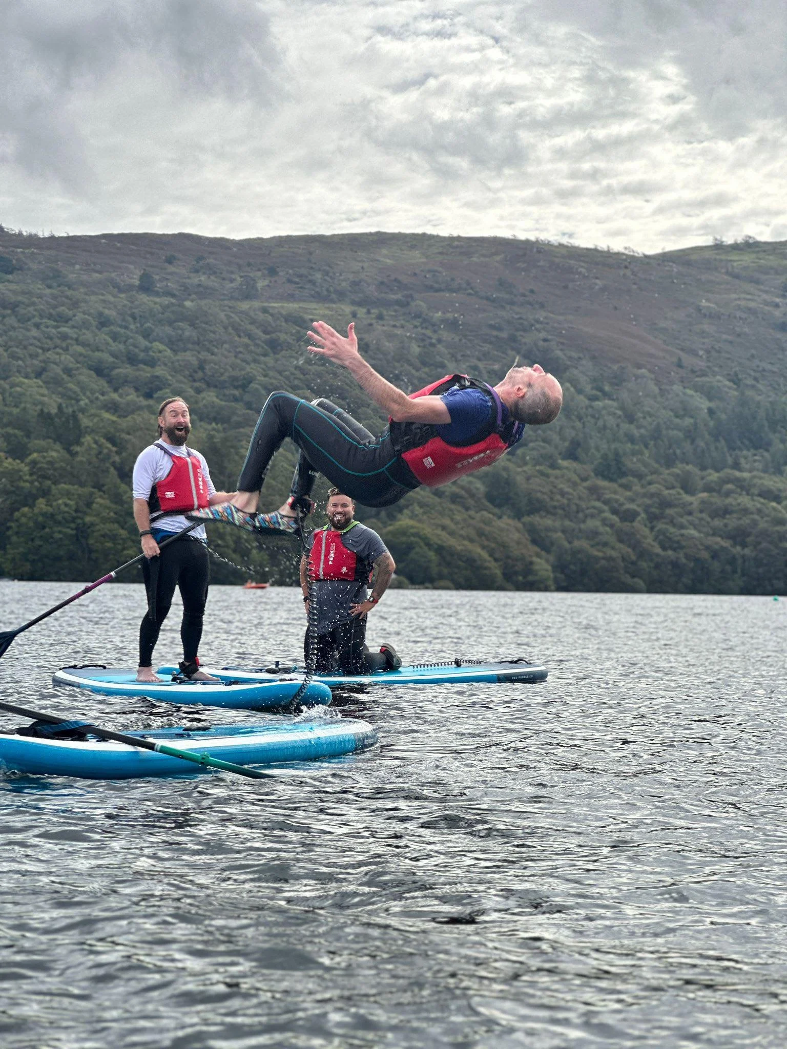Three men on paddleboards on a lake, with one man jumping into the water, surrounded by green hills and overcast sky.
