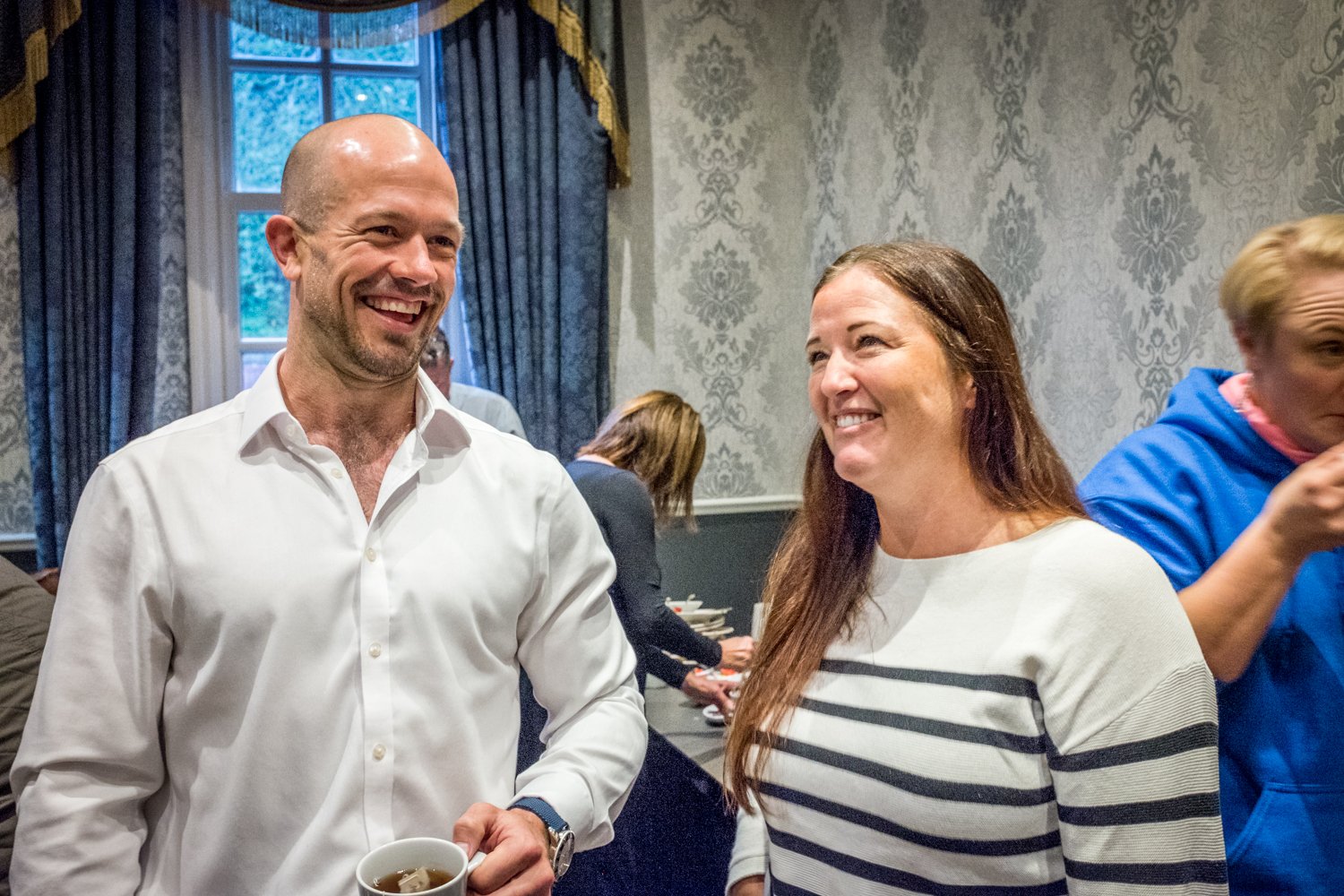 Two people smiling and engaging in a conversation at an indoor gathering. The man on the left is holding a white mug, and a woman with long brown hair is on the right. The background features patterned wallpaper and other people.