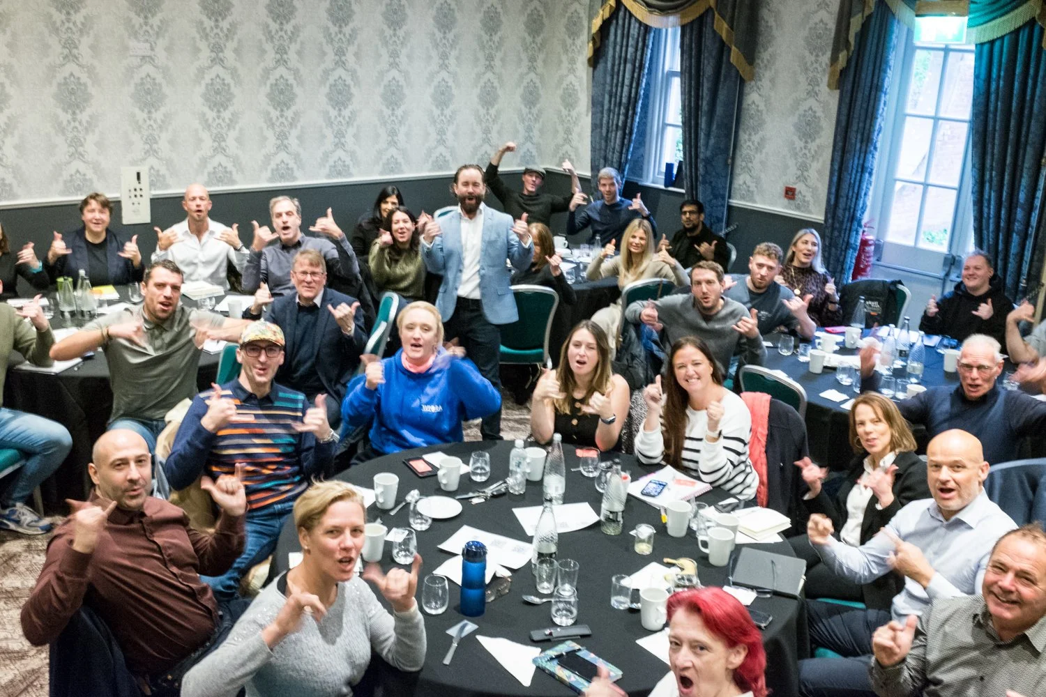 A large group of people gathered around several round tables in a banquet-style room, posing for a group photo and making thumbs-up gestures. The room has patterned wallpaper, large windows with curtains, and a fire extinguisher visible near the window.