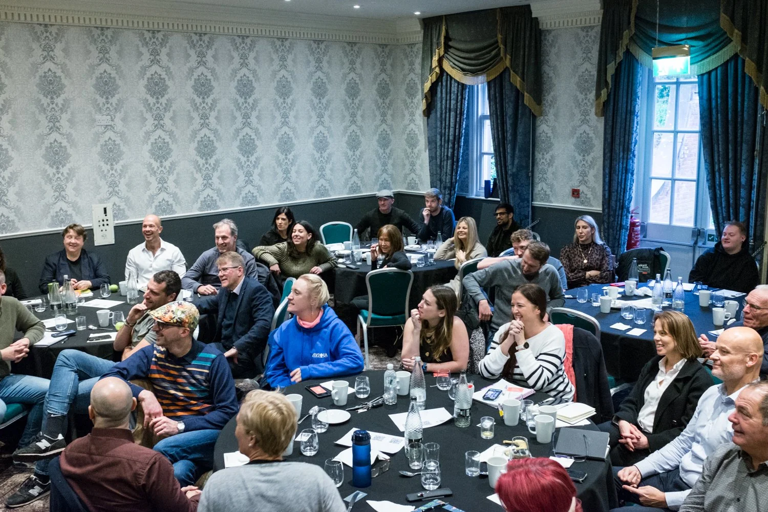 A group of people sitting around tables in a conference room, smiling and engaging during a presentation or discussion.