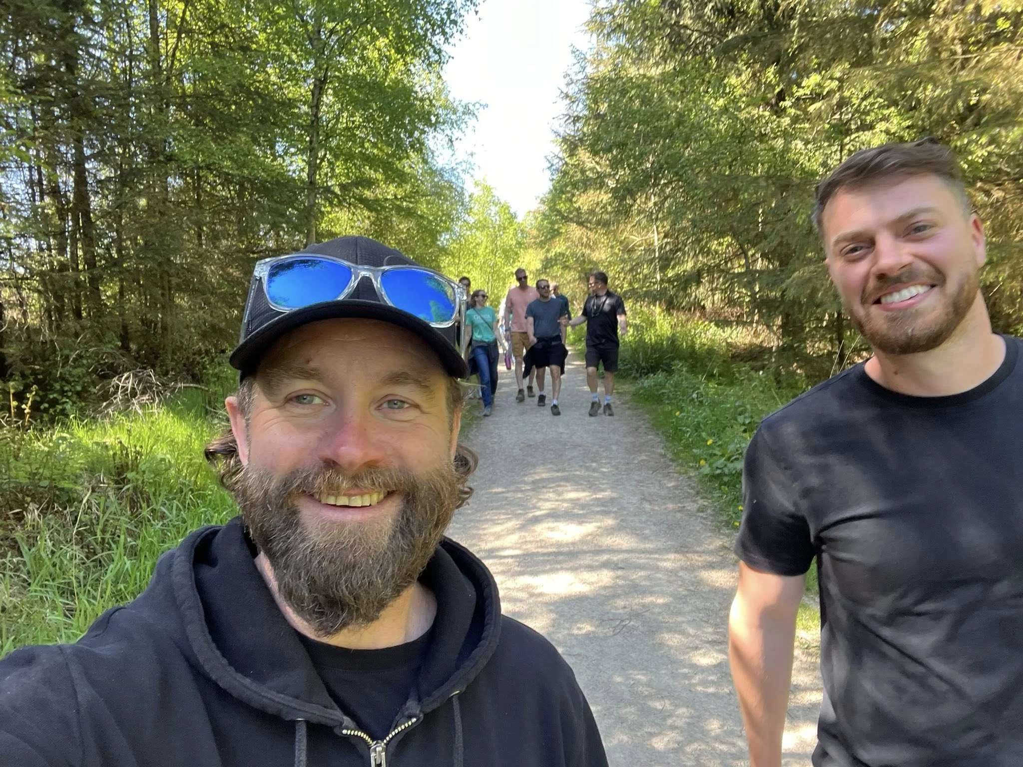 Two smiling men taking a selfie on a forest trail with a group of people walking behind them.