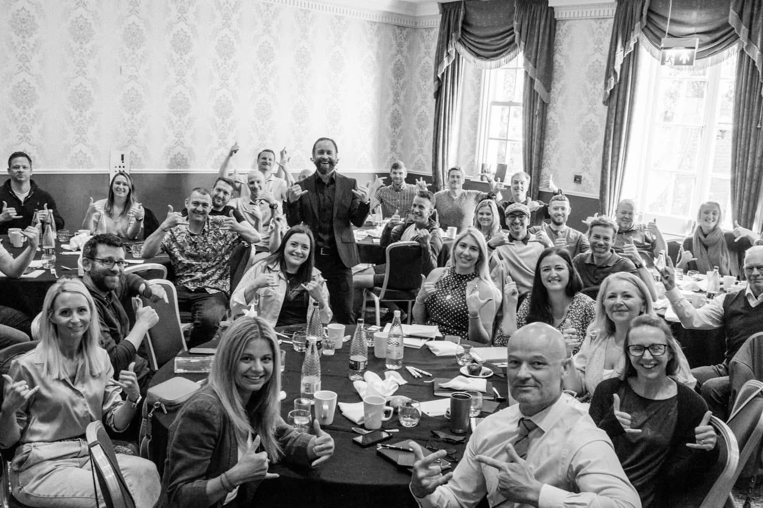A large group of people, mostly smiling and making hand gestures, gathered in a bright, ornate room for a celebration or meeting. They are seated around tables with drinks and tableware. A man in a suit stands in the center. The room has large windows with curtains and patterned wallpaper.
