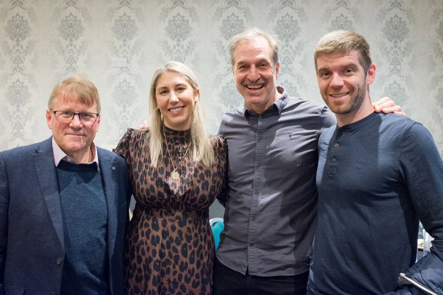 Four people standing together indoors in front of a patterned wallpaper, smiling at the camera.