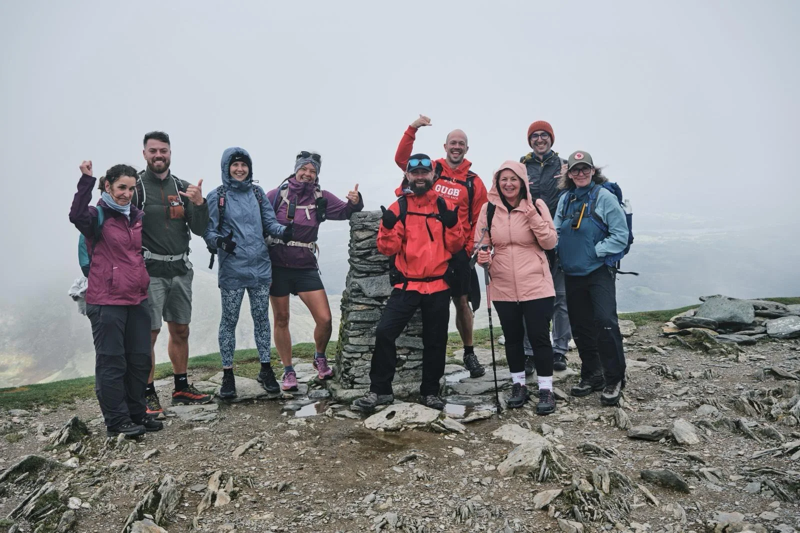 Group of ten hikers, dressed in outdoor gear, standing on a rocky mountain summit with a foggy background, celebrating their achievement.