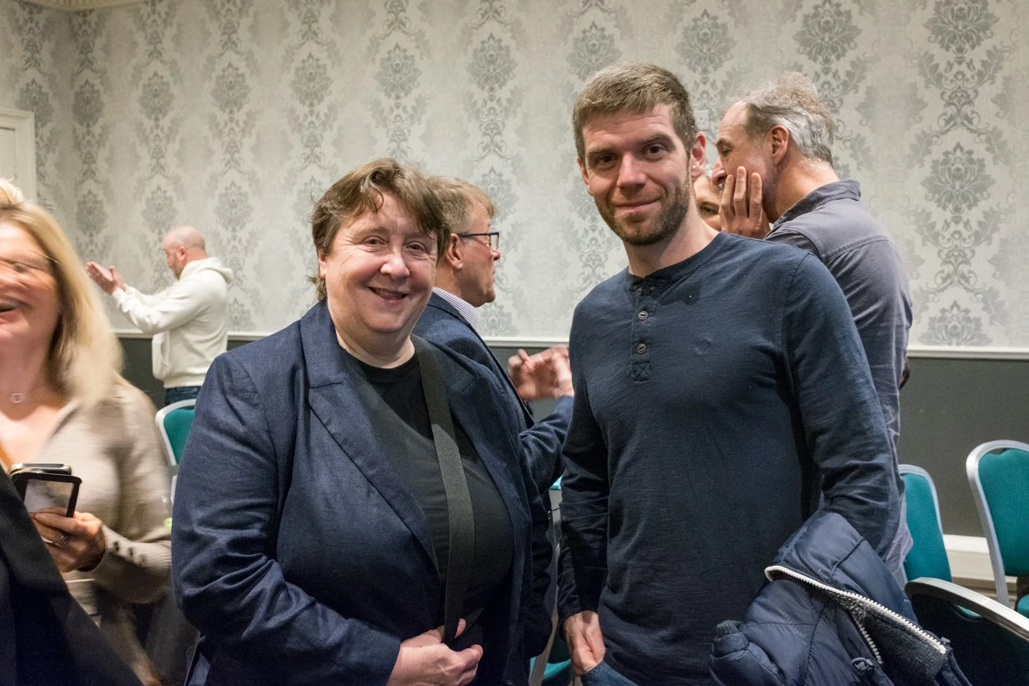 Two men standing together in a room, smiling at the camera, with people engaged in conversations in the background.