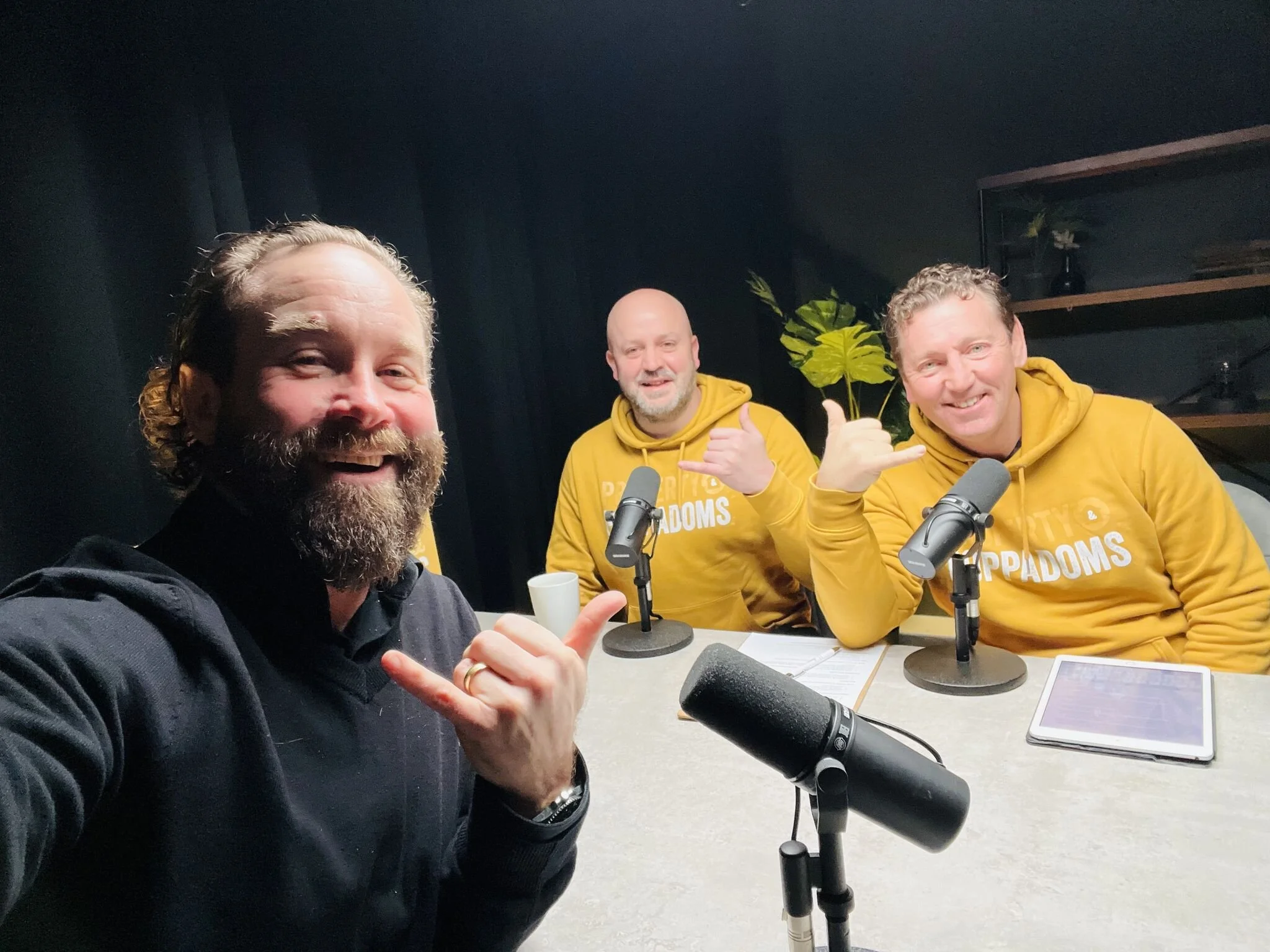 Three smiling men sitting at a table with microphones, two wearing yellow hoodies with 'PROPAGANDUMS' printed on them, in a dark room with shelves and a plant in the background.