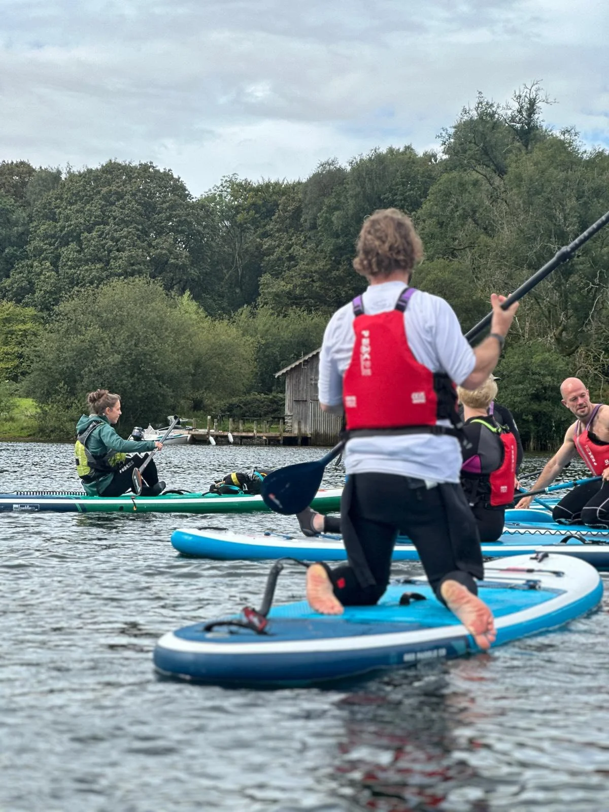 Group of people paddleboarding on a lake with trees and a small boathouse in the background.