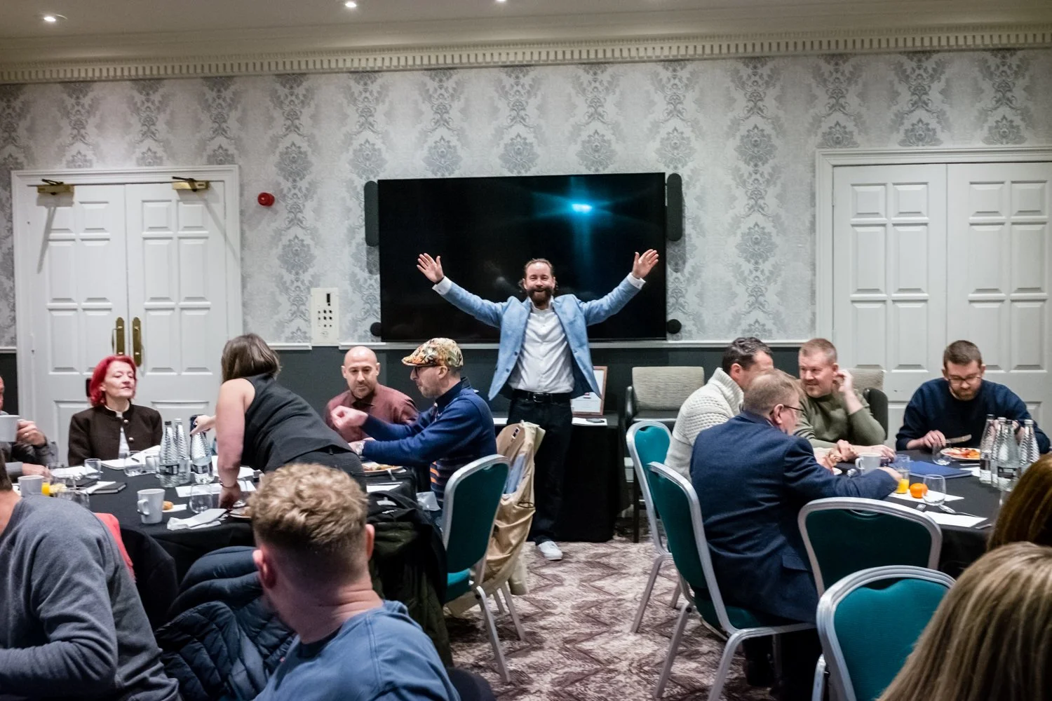 People sitting around tables in a conference room, some eating and drinking, while a man at the front with arms outstretched smiles at the camera, with a large black screen behind him.