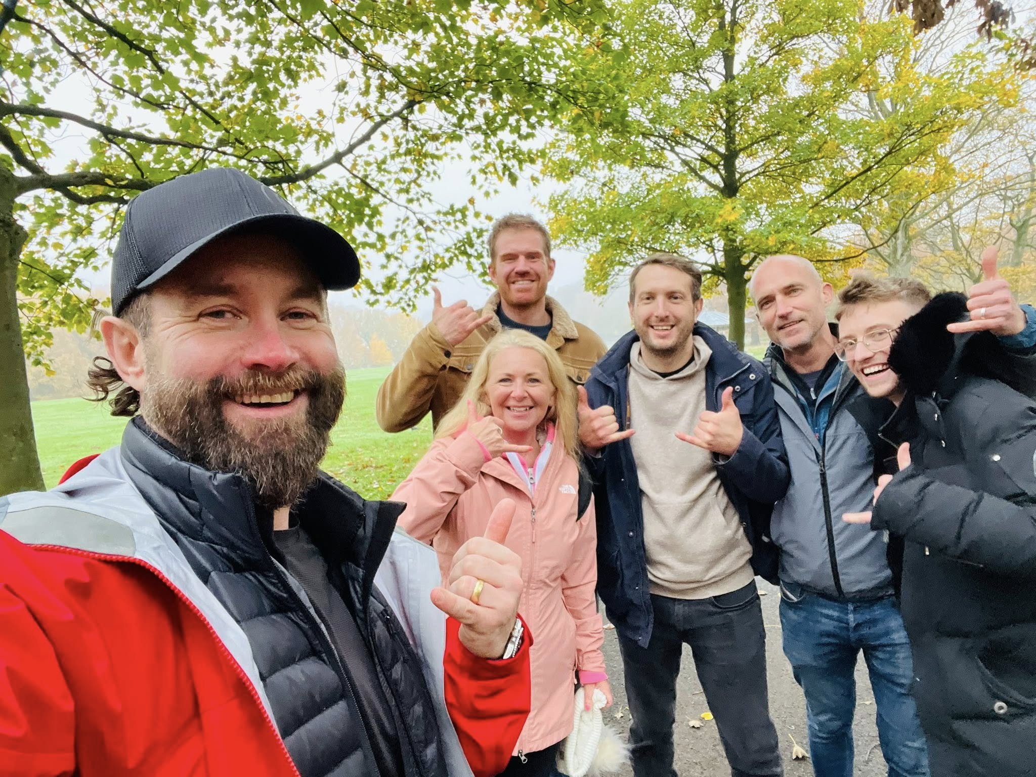 Group of seven people outdoors in a park with trees showing fall foliage, smiling, and making gestures for fun.