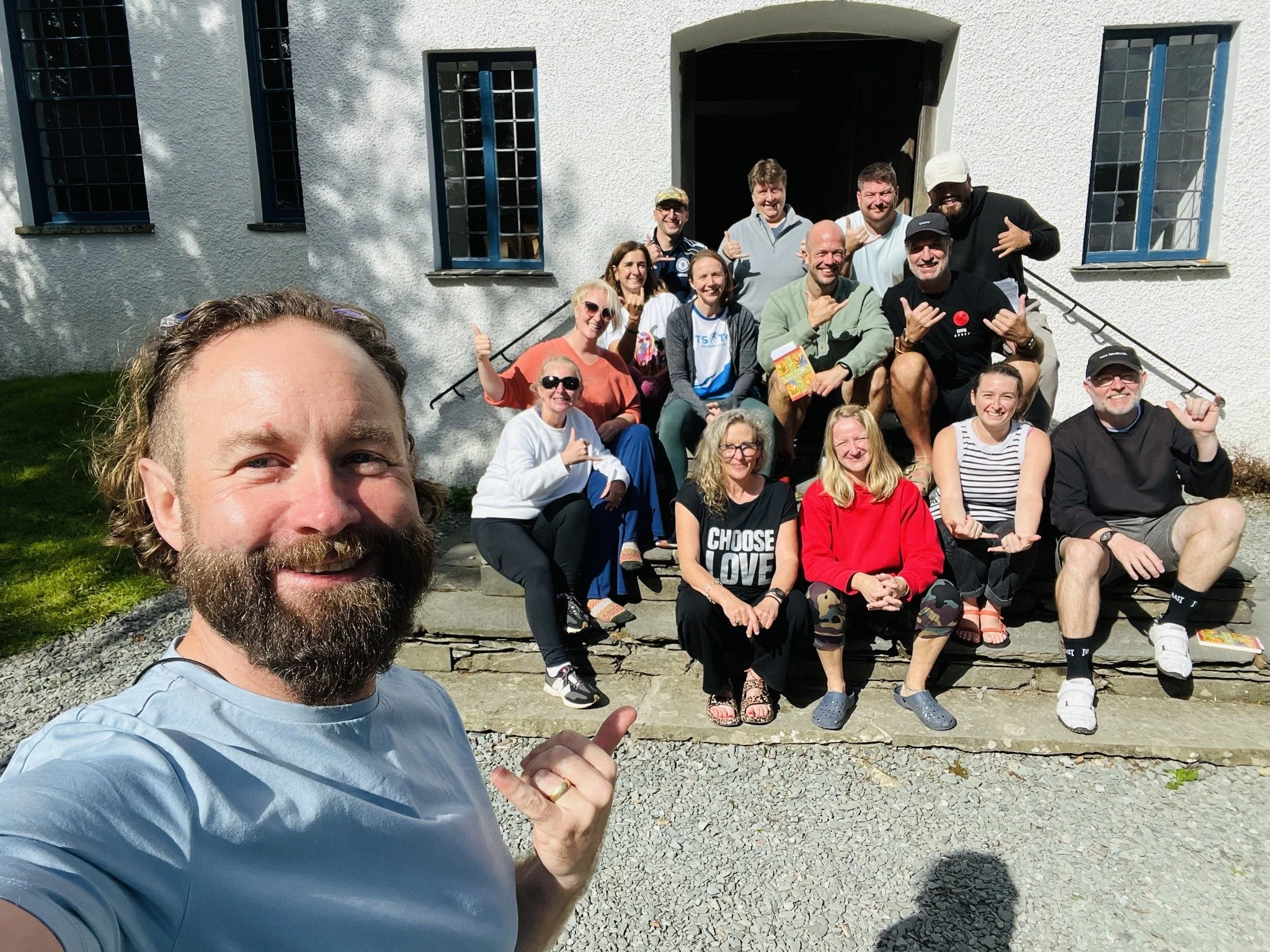 A man with a beard taking a selfie with a large group of people sitting and standing outside a white building with blue windows. Everyone is smiling and making hand gestures, enjoying a sunny day.