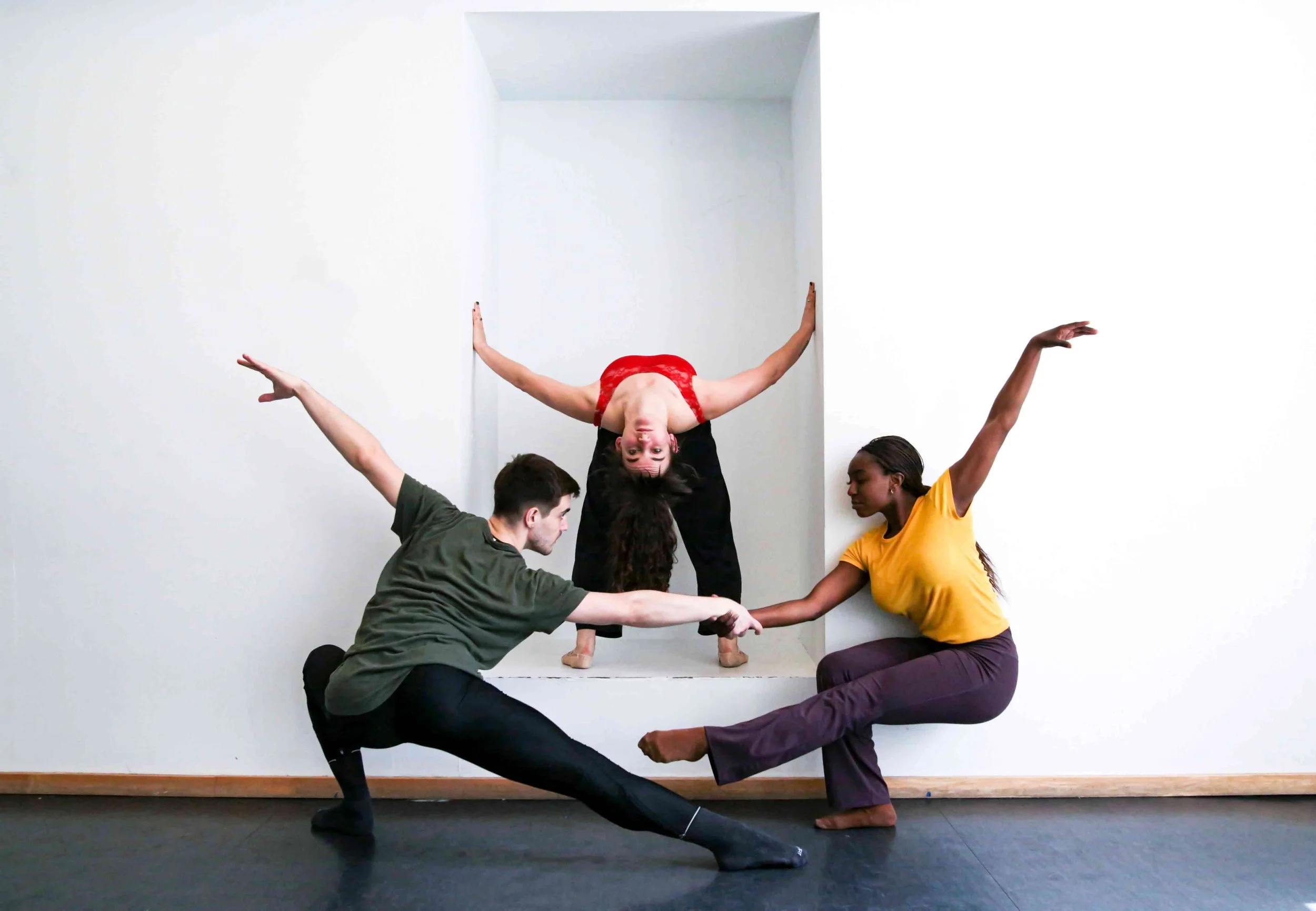 Three Shades Dance Theater dancers pose in a room with white walls and black flooring. One dancer is upside down in a wall-supported handstand with arms extended and wearing a red top. The other two dancers are on the floor.