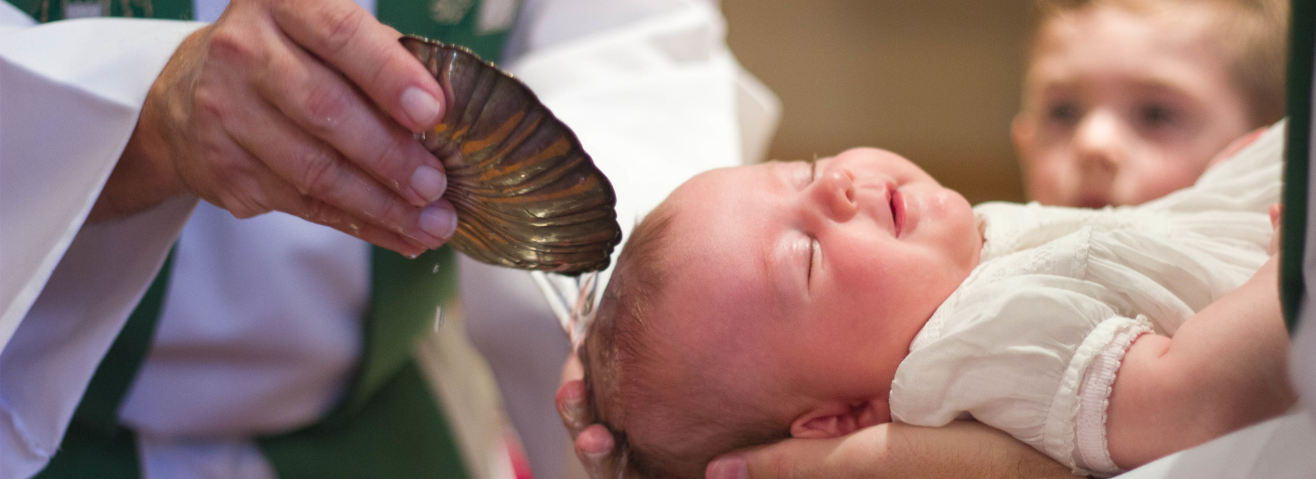 Person holding a baby over a water basin during a baptism ceremony, with another child watching in the background.