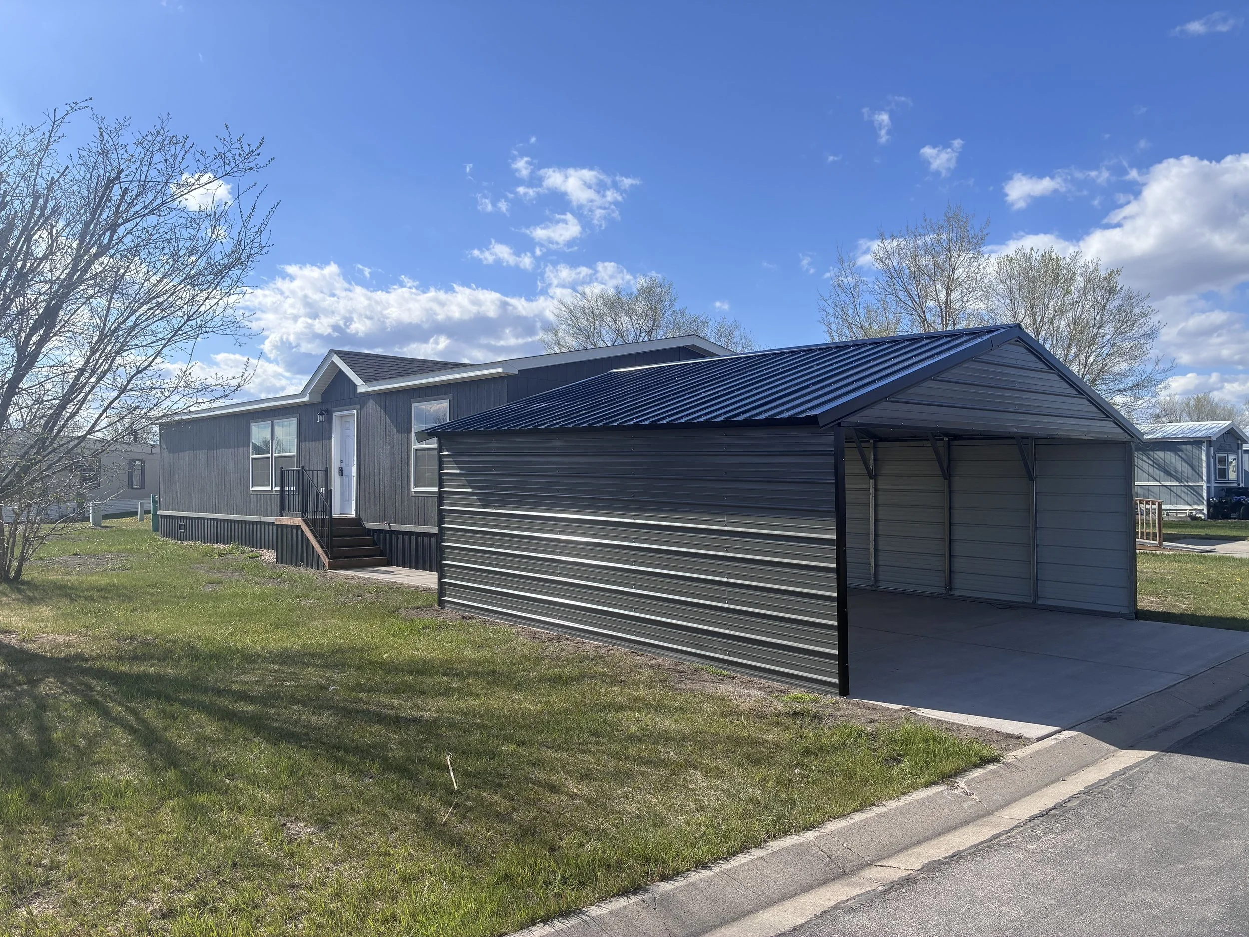 A black manufactured home on a grassy lot with a small set of stairs leading to a white door. Multiple windows on the front side and trees in the background under a blue sky.