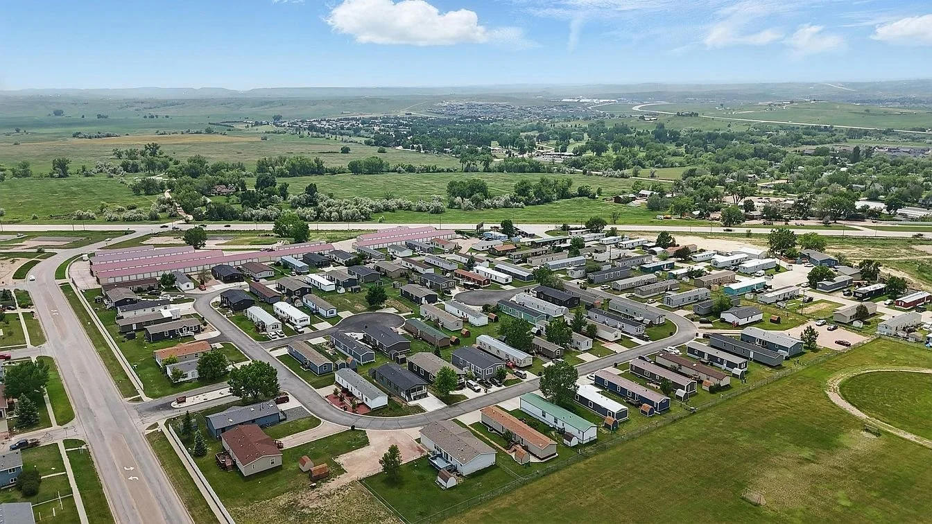 Aerial view of a suburban mobile home park with multiple manufactured homes and RVs, surrounded by green fields and open countryside under a partly cloudy sky.