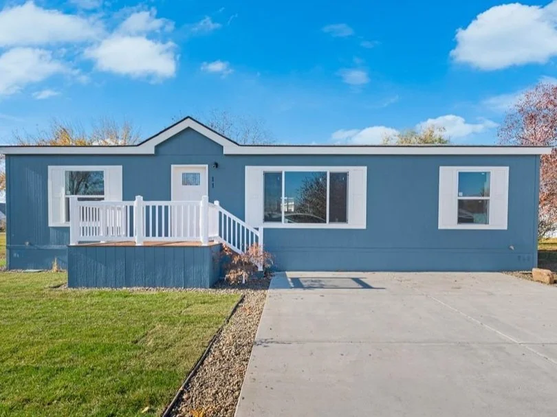 Blue single-story house with white trim, a small front porch with white railing, and large windows, set in a grassy yard with a concrete driveway, under a partly cloudy sky.