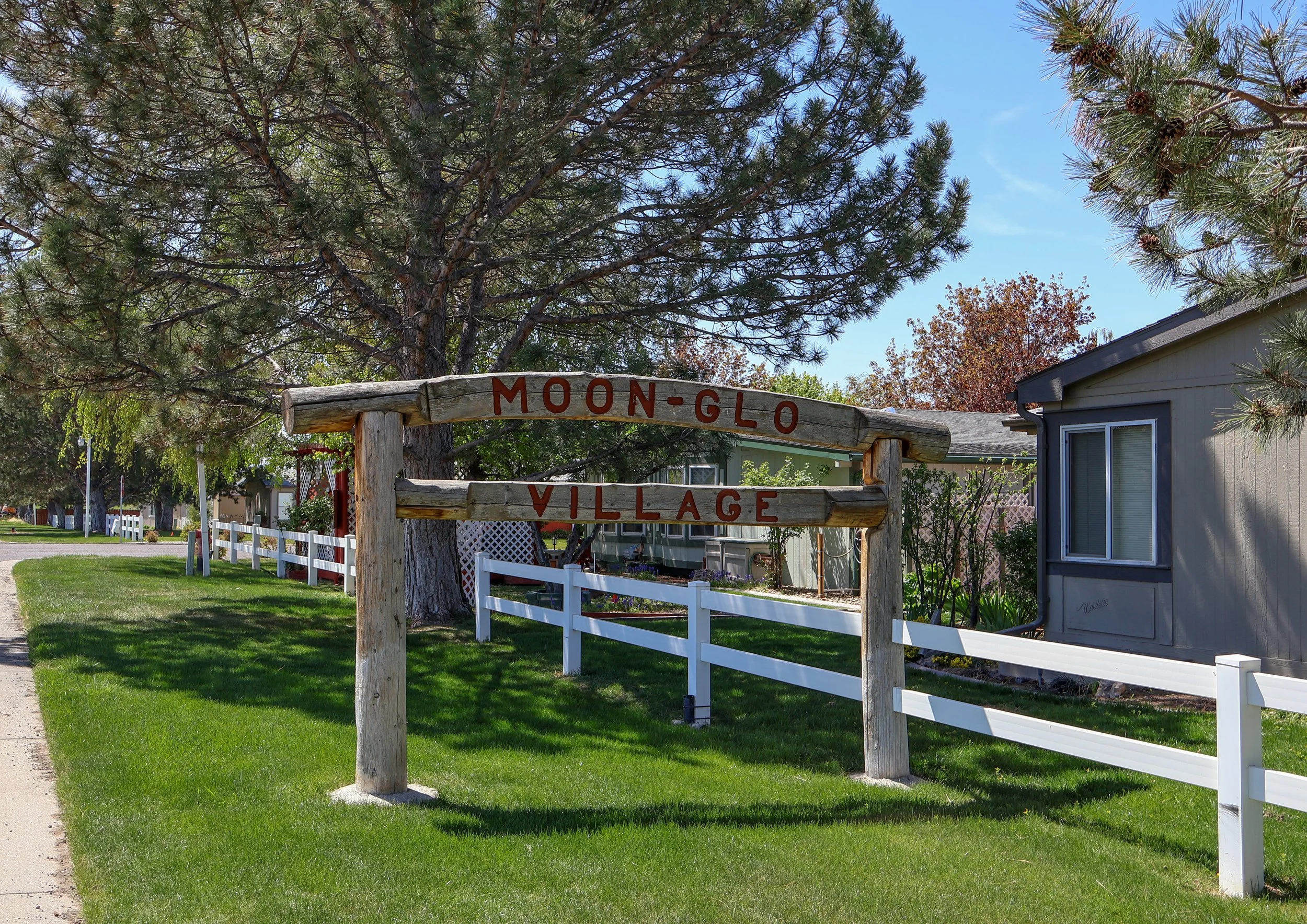 Sign reading 'Moon-Glo Village' located at the entrance of a residential area with a white fence, green grass, and trees in the background.