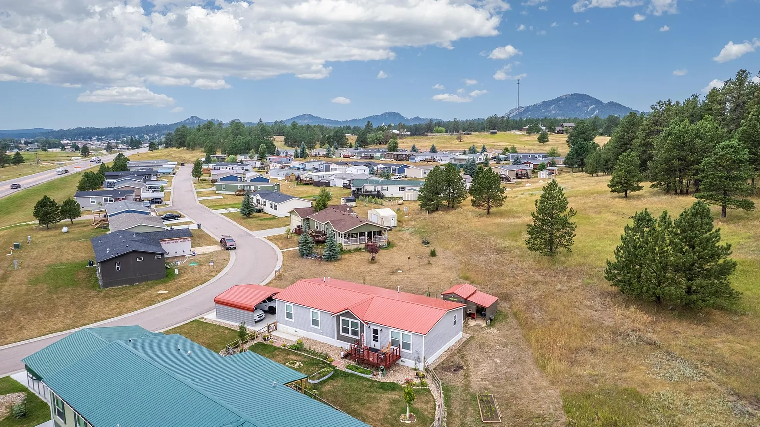 Aerial view of a suburban neighborhood with homes, a curved street, green lawns, trees, and distant mountains under a partly cloudy sky.