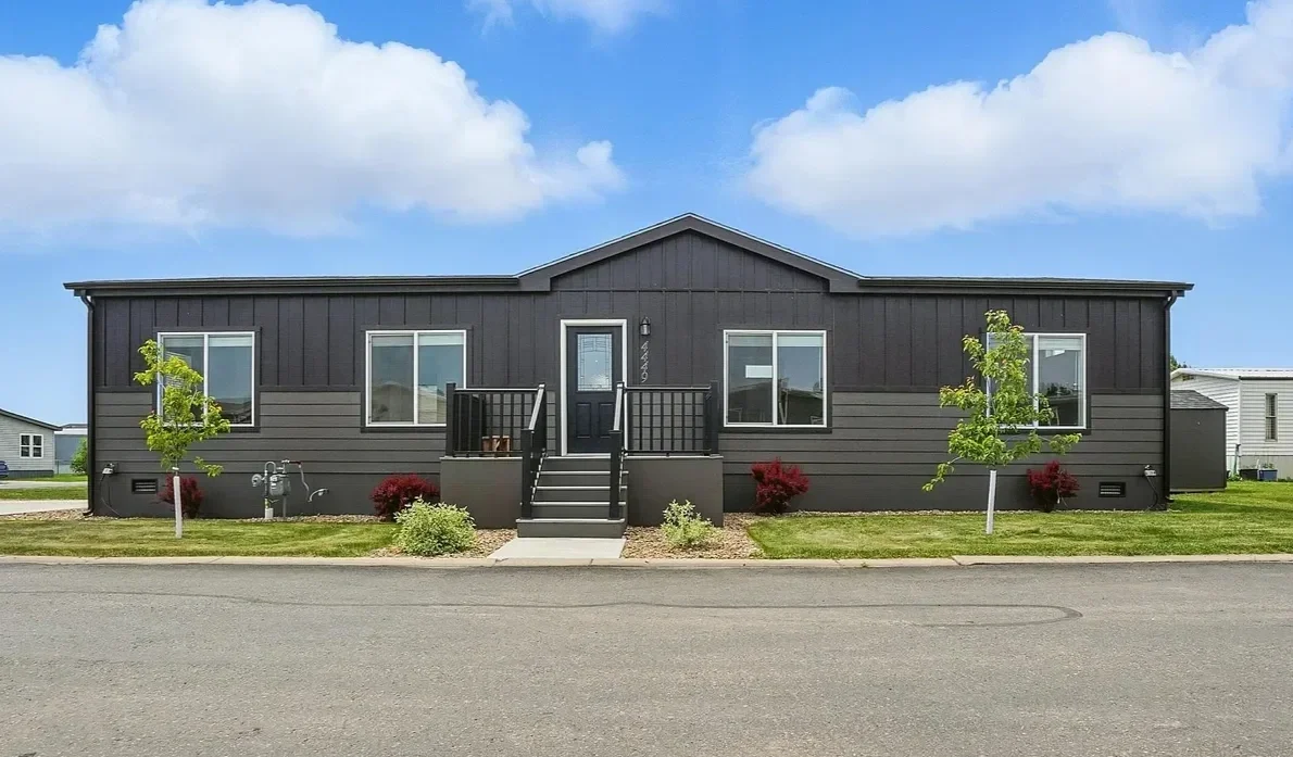 A single-story, dark gray manufactured home with a small front outdoor staircase, two small trees, and landscaped bushes on a suburban street under a partly cloudy blue sky.