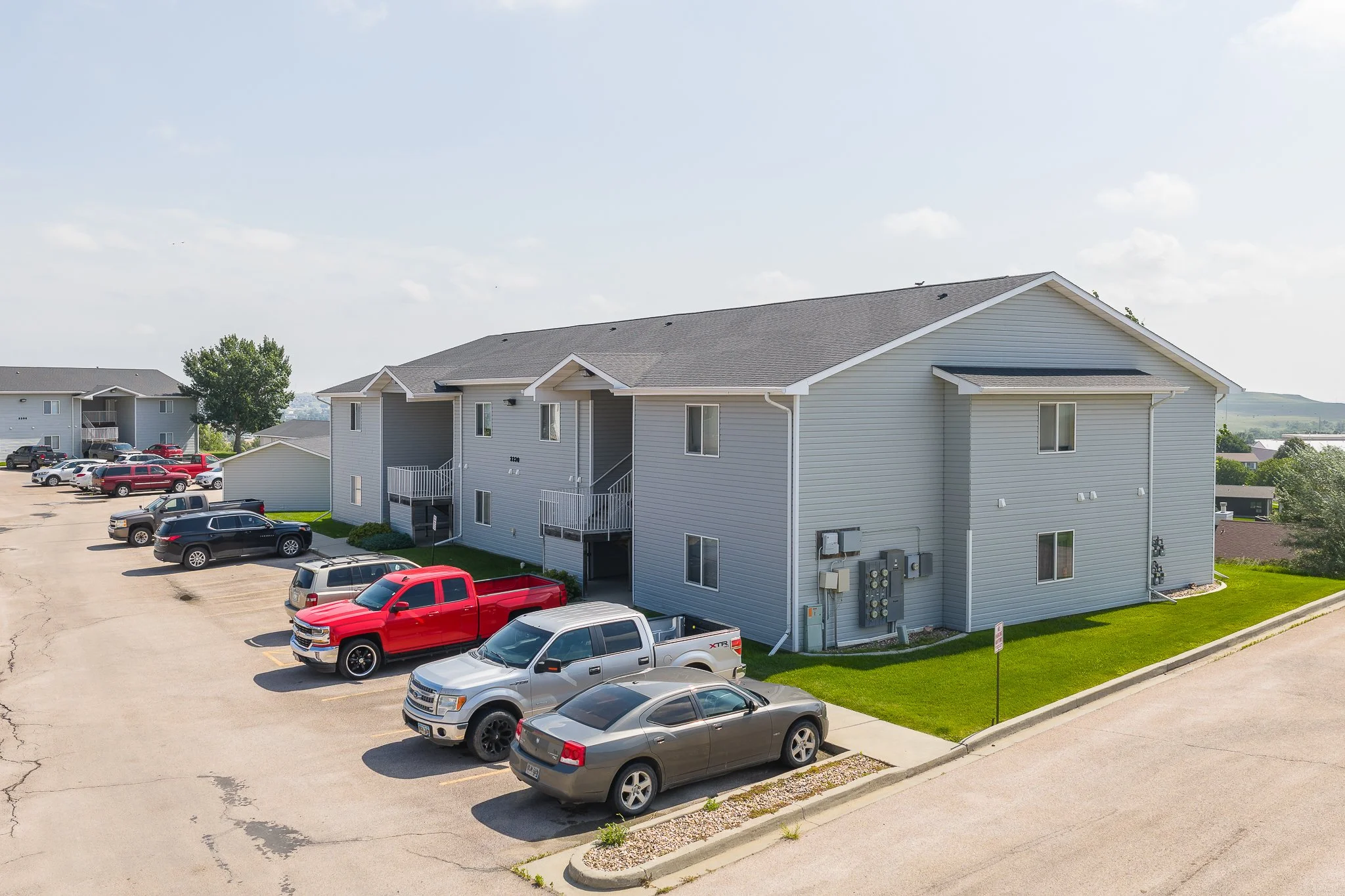 Apartment building with parking lot and several parked cars, including trucks, SUVs, and sedans, under a partly cloudy sky.