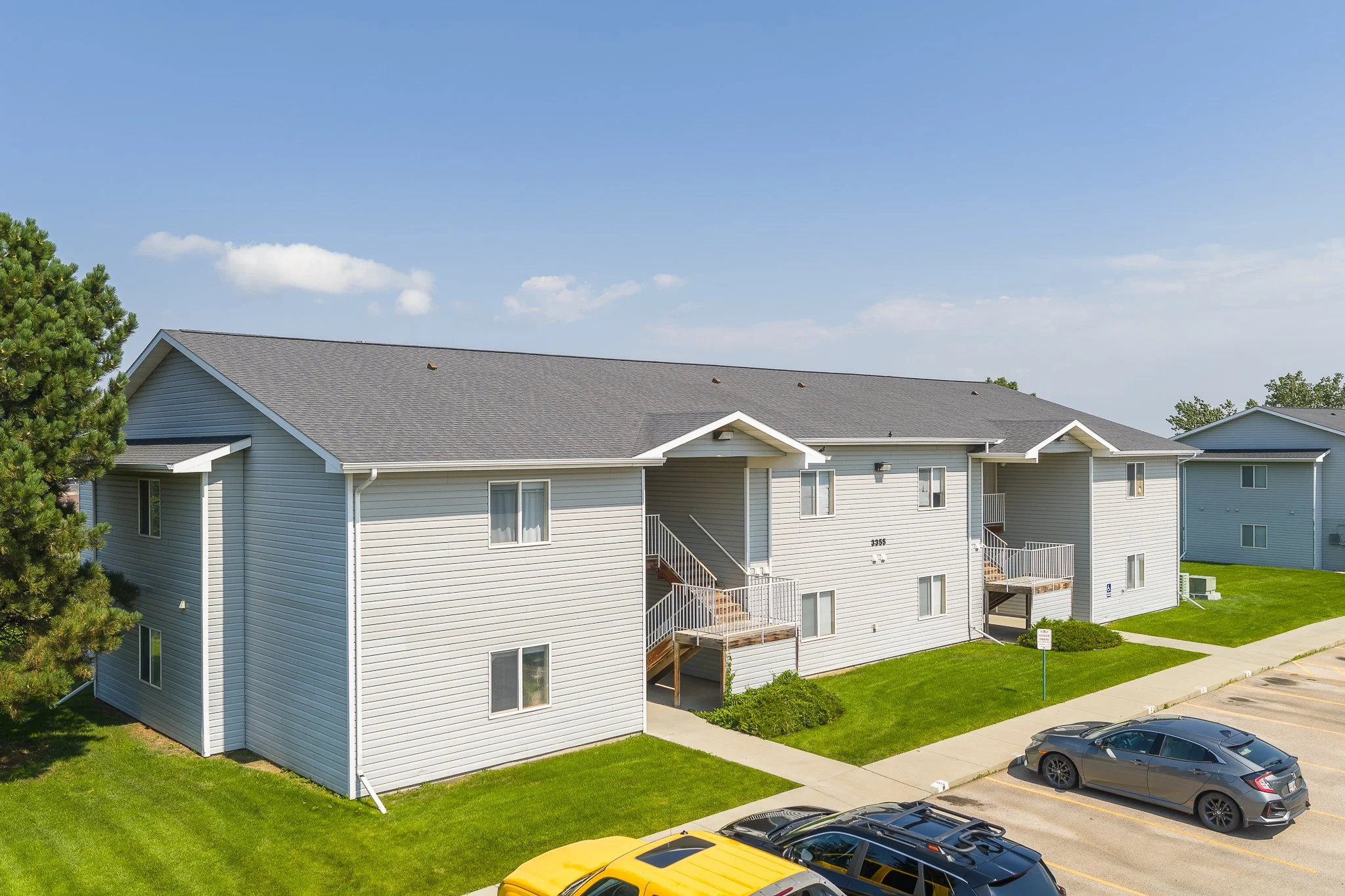 Exterior view of a multi-unit residential building with gray siding, multiple windows, small balconies, a grassy lawn, and parked cars in a parking lot.