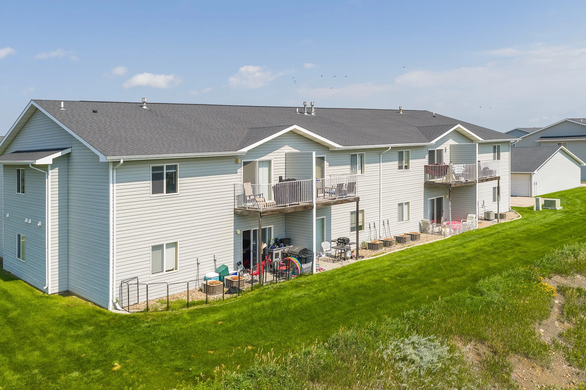 Multi-family residential building with balconies, outdoor furniture, and toys on a grassy hill under a blue sky.