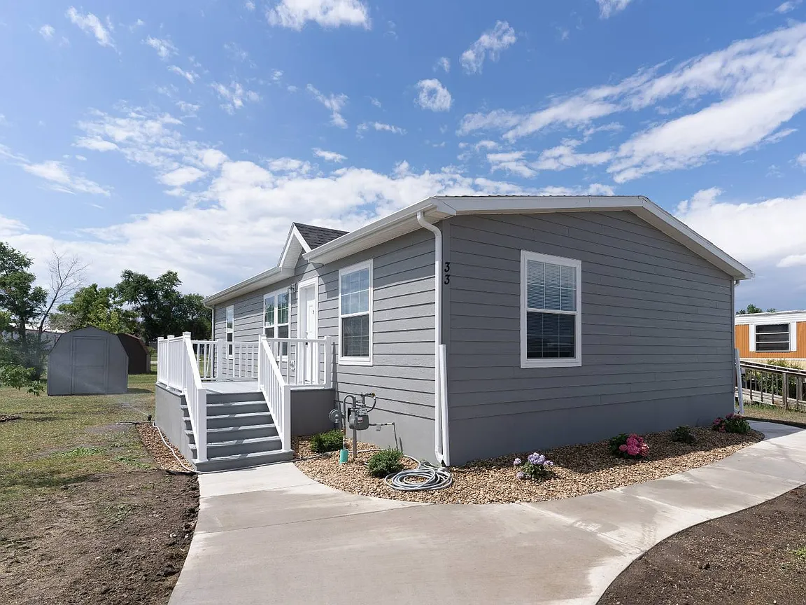 A gray house with white trim, a small porch with stairs, and a curved sidewalk leading to the front yard, with bushes and a garden bed along the foundation. The sky is partly cloudy.