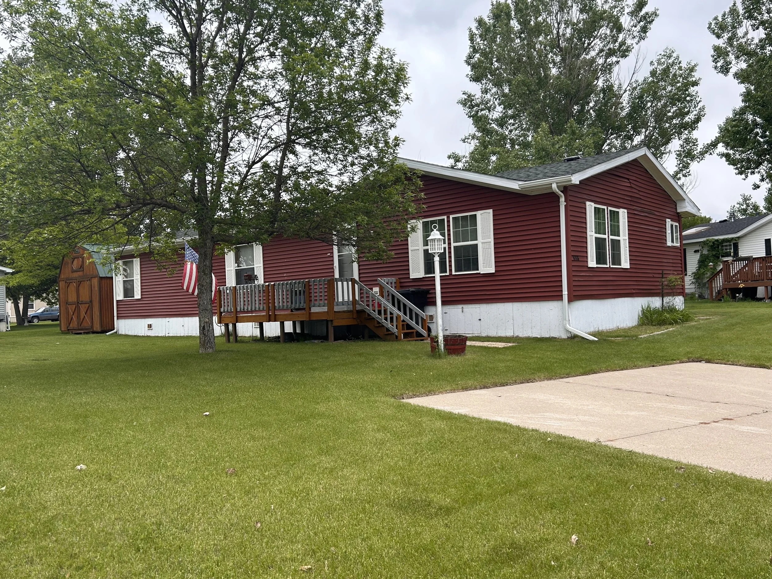 A red house with white window shutters and a small front deck, surrounded by a green lawn and trees, with an American flag hanging on the side.