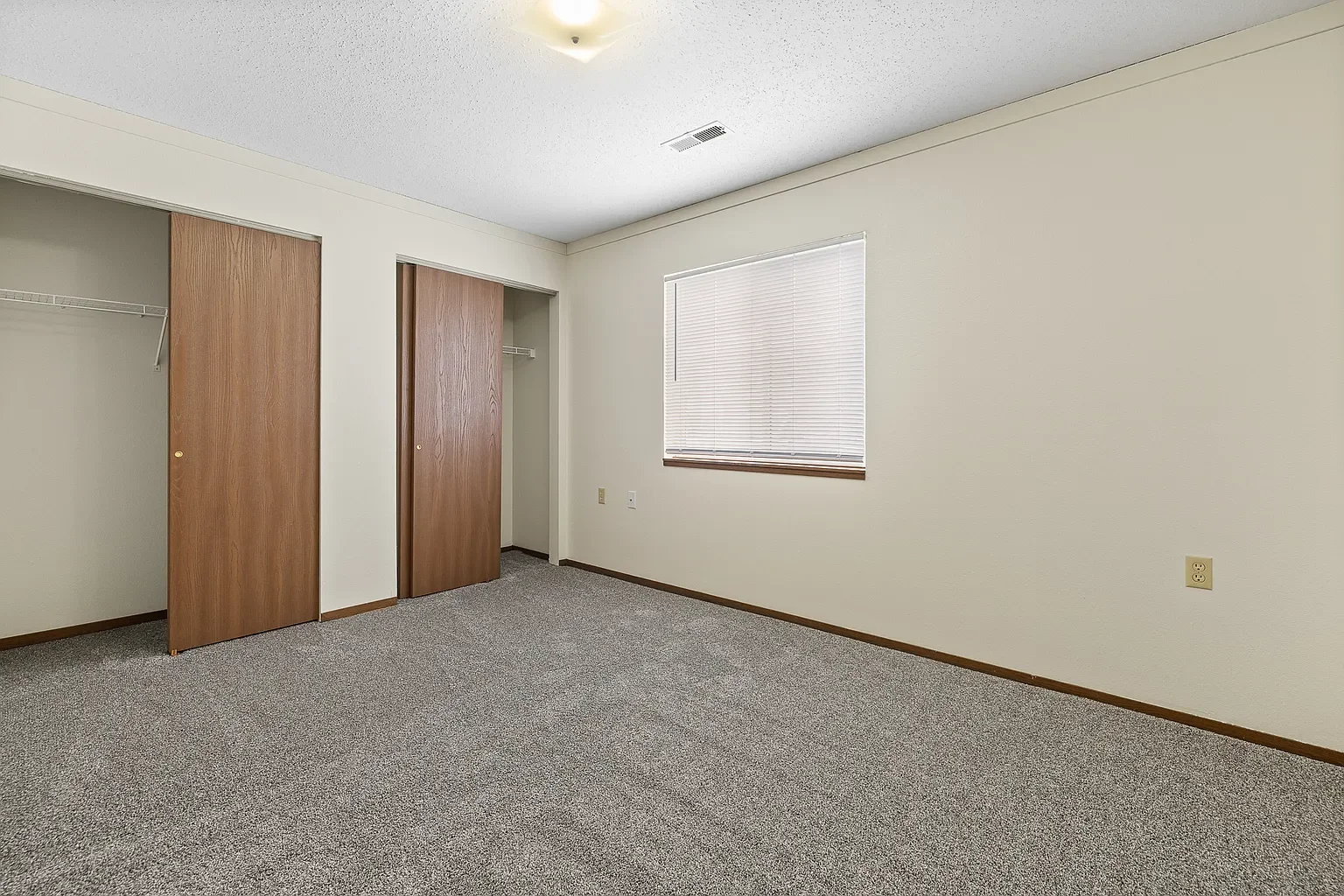 Empty bedroom with beige walls, carpeted floor, a window with blinds, and an open closet with wooden sliding doors.