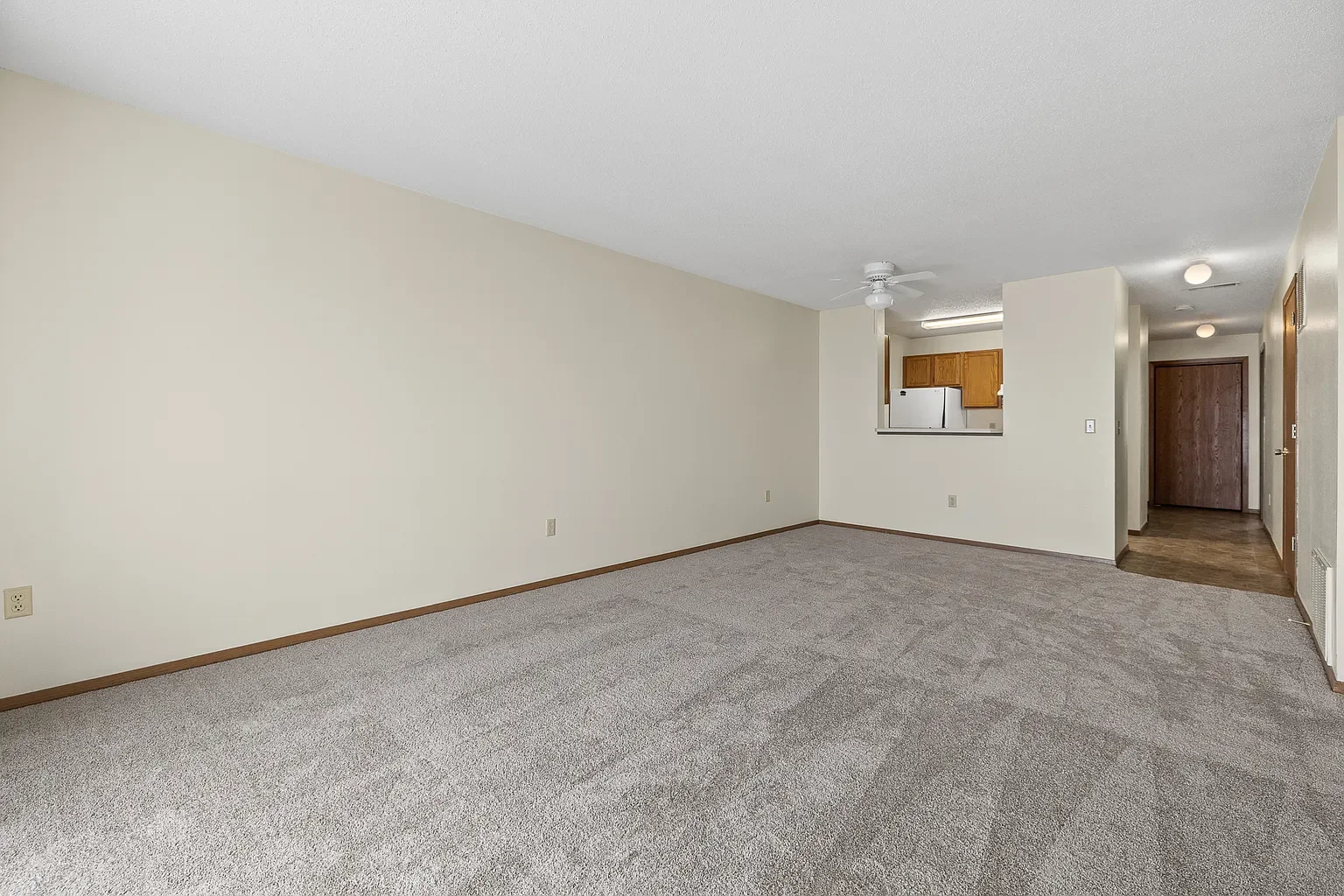 Empty living room with beige walls, gray carpet, and view into kitchen with wooden cabinets and white refrigerator.