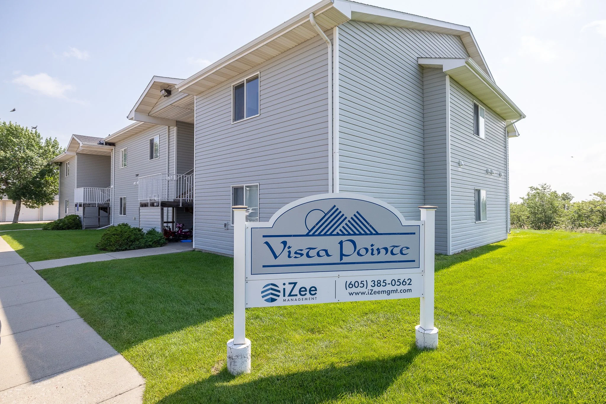 A multi-story residential building with light gray siding, small balconies, and white trim. In front, there is a sign reading 'Vista Pointe' with the iZee Management logo, contact number, and website. The building is surrounded by a well-maintained g