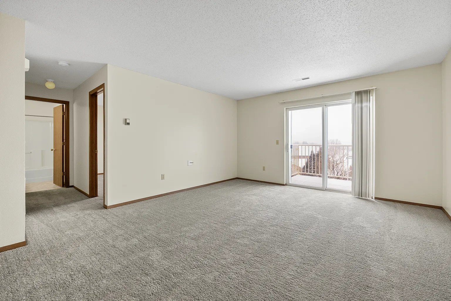Empty living room with beige carpet, white walls, large glass sliding door leading to a balcony, and a hallway with doors leading to bathroom and bedroom.