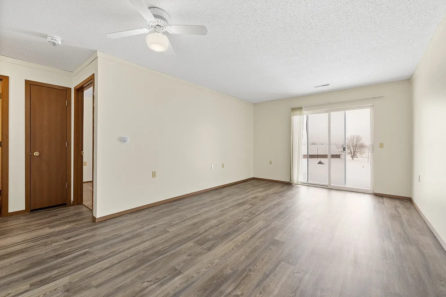 Empty living room with wood flooring, white walls, a ceiling fan, large sliding glass door, and a window showing a snowy outdoor scene.
