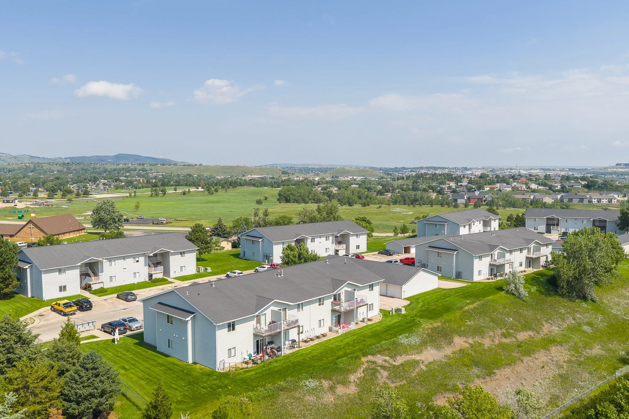 Aerial view of multiple two-story apartment buildings with green lawns, parking lots, and trees in a suburban area with open fields and hills in the background.