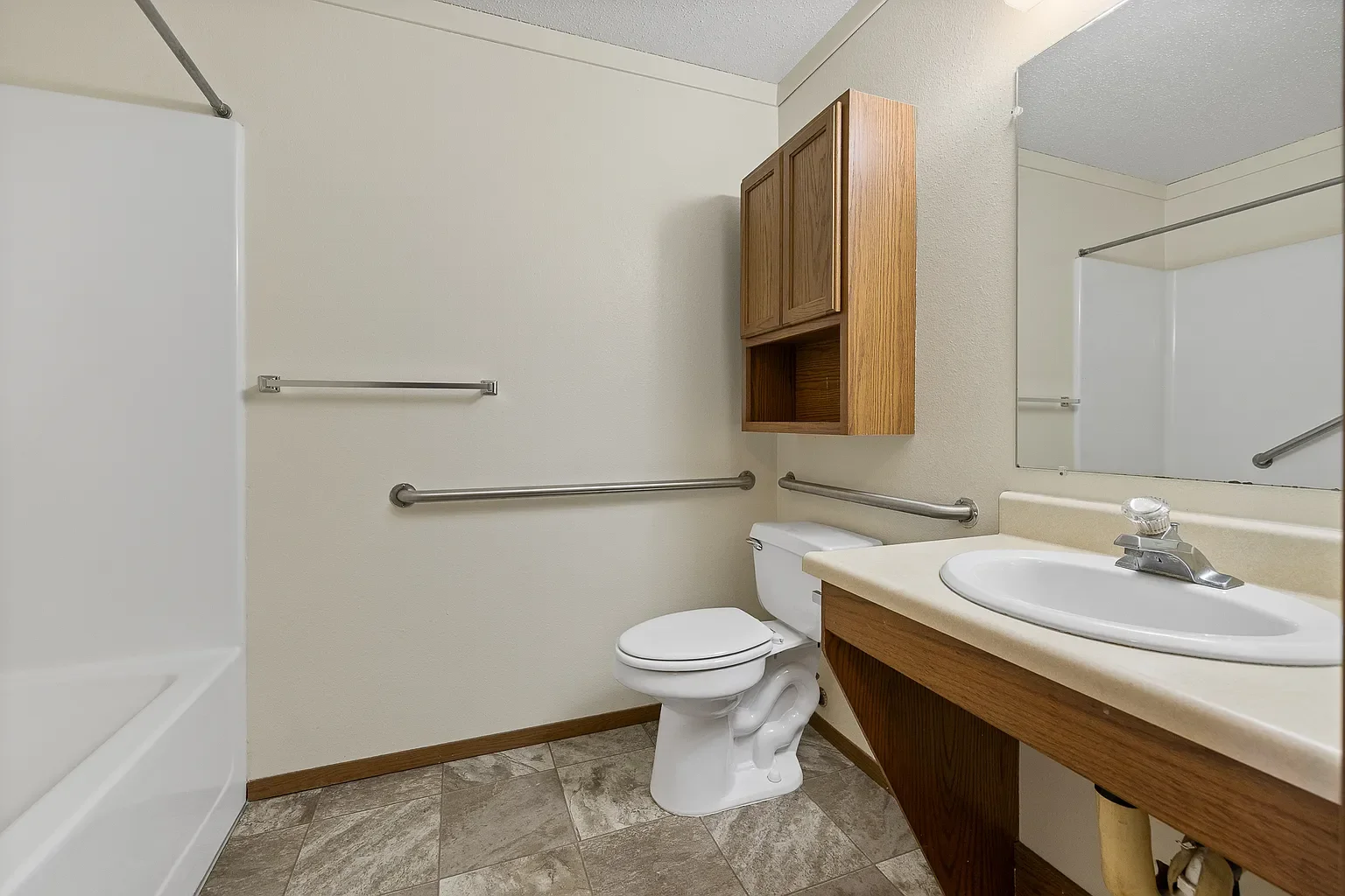 A plain bathroom with a white bathtub, toilet, wooden wall-mounted cabinet, beige countertop with a sink, large mirror, and beige tiled flooring.