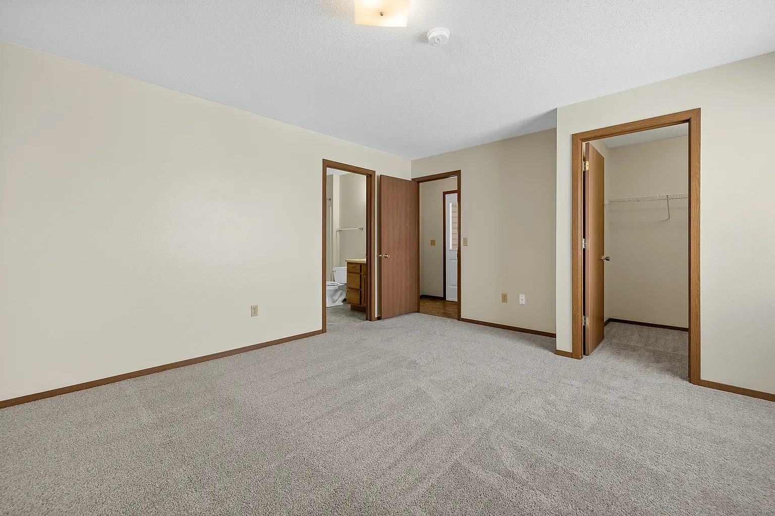 Empty bedroom with beige walls, carpeted floor, and two open closet doors, one leading to a walk-in closet and the other to an adjacent room with a bathroom and a hallway visible in the background.