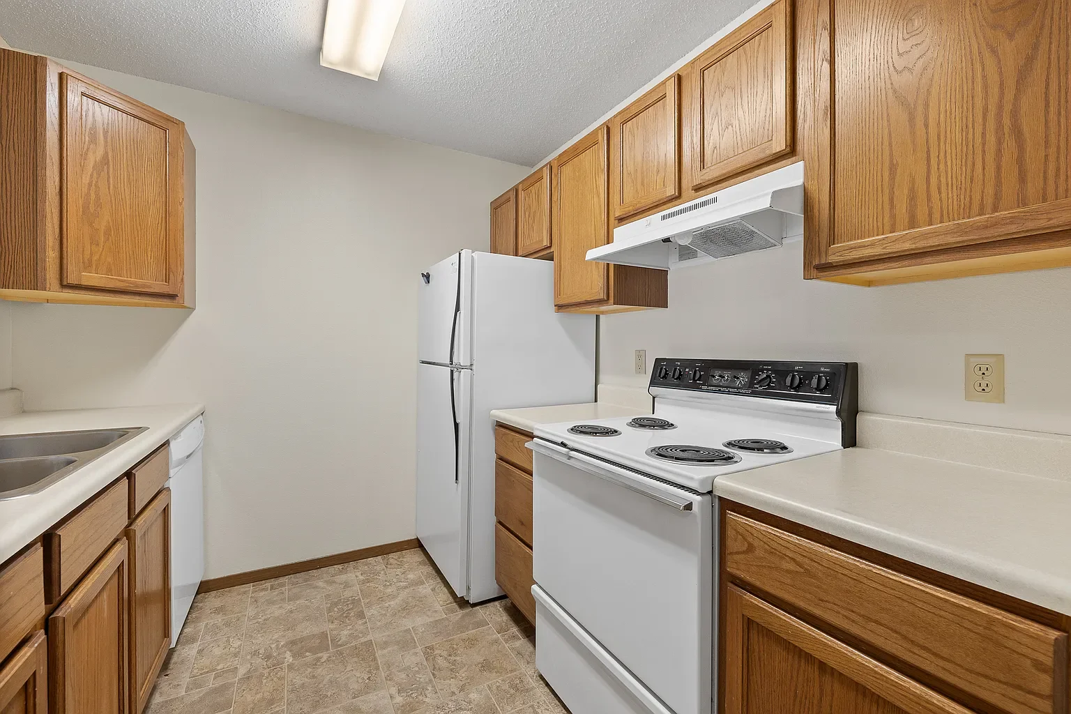 Kitchen with wooden cabinets, white electric stove, refrigerator, and double sink, beige countertops, tan tiled floor, ceiling light, and electrical outlets.