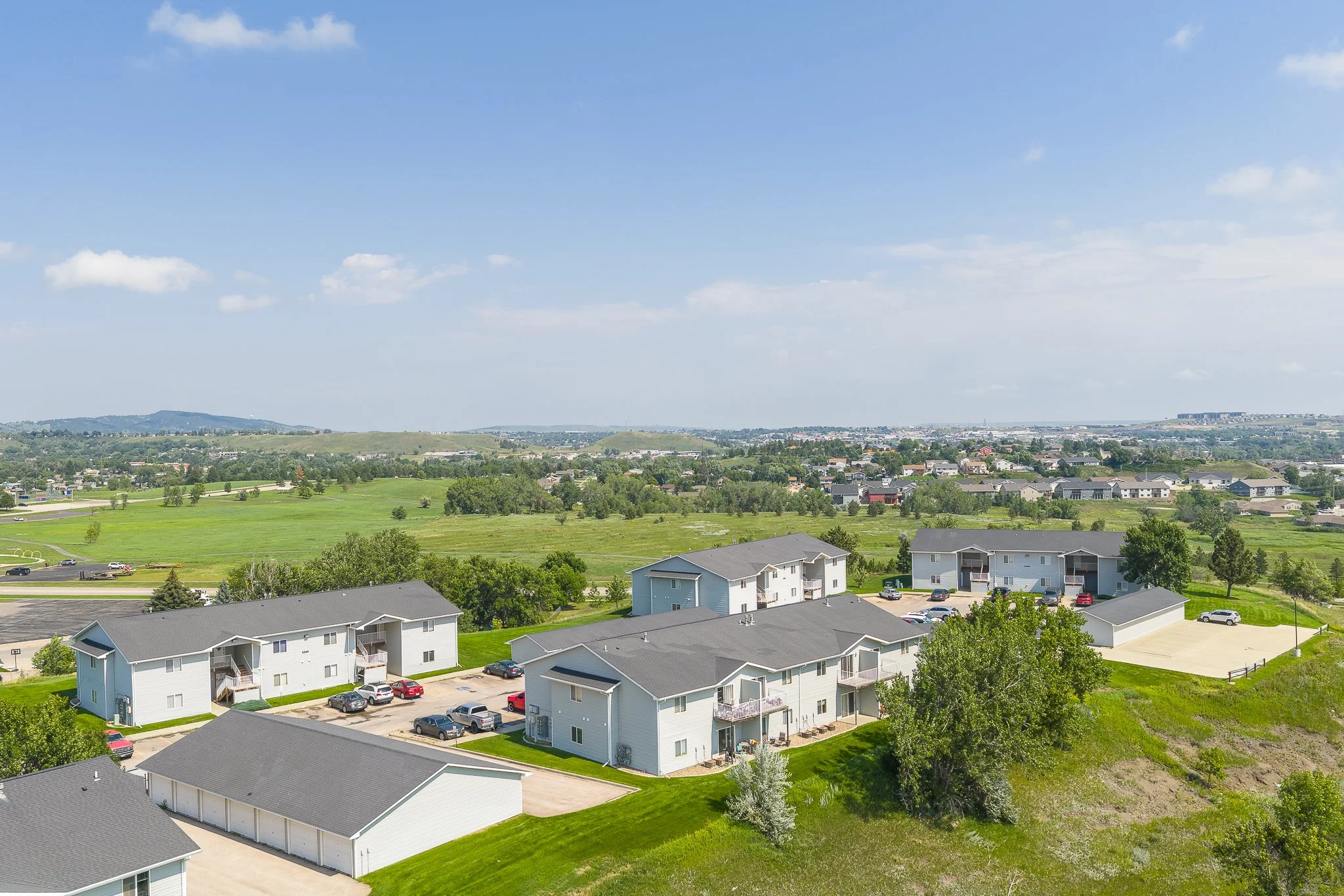 Aerial view of a residential neighborhood with multiple white apartment buildings, parking lots, green lawns, and trees, under a partly cloudy sky.