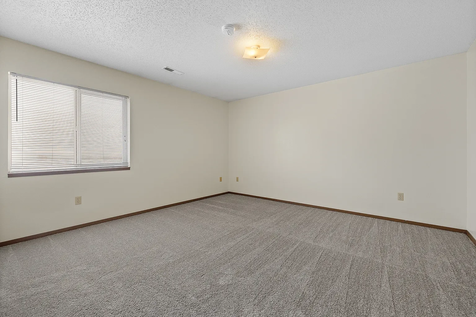 Empty room with beige walls, carpeted floor, window with blinds, ceiling light, and electrical outlets.