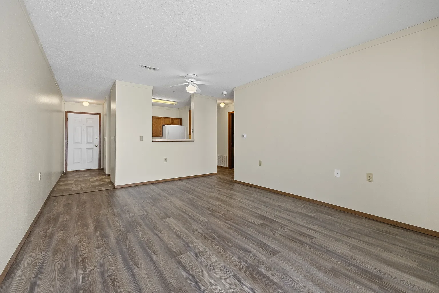 Empty living room with wood laminate flooring, beige walls, a ceiling fan, and view into the kitchen with wooden cabinets and white appliances.