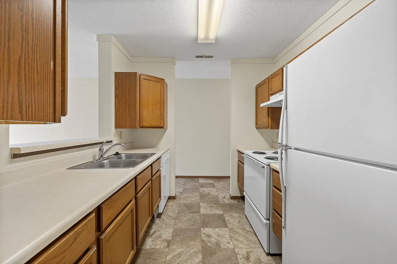 Kitchen with wooden cabinets, white appliances including a refrigerator, stove, and dishwasher, double sink, and tiled floor.