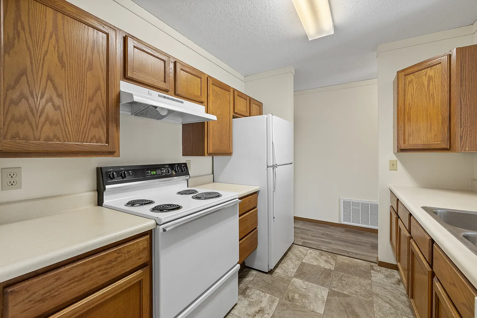 Kitchen with wooden cabinets, white stove, white refrigerator, beige countertops, and double sink. Tiled floor and fluorescent ceiling light.