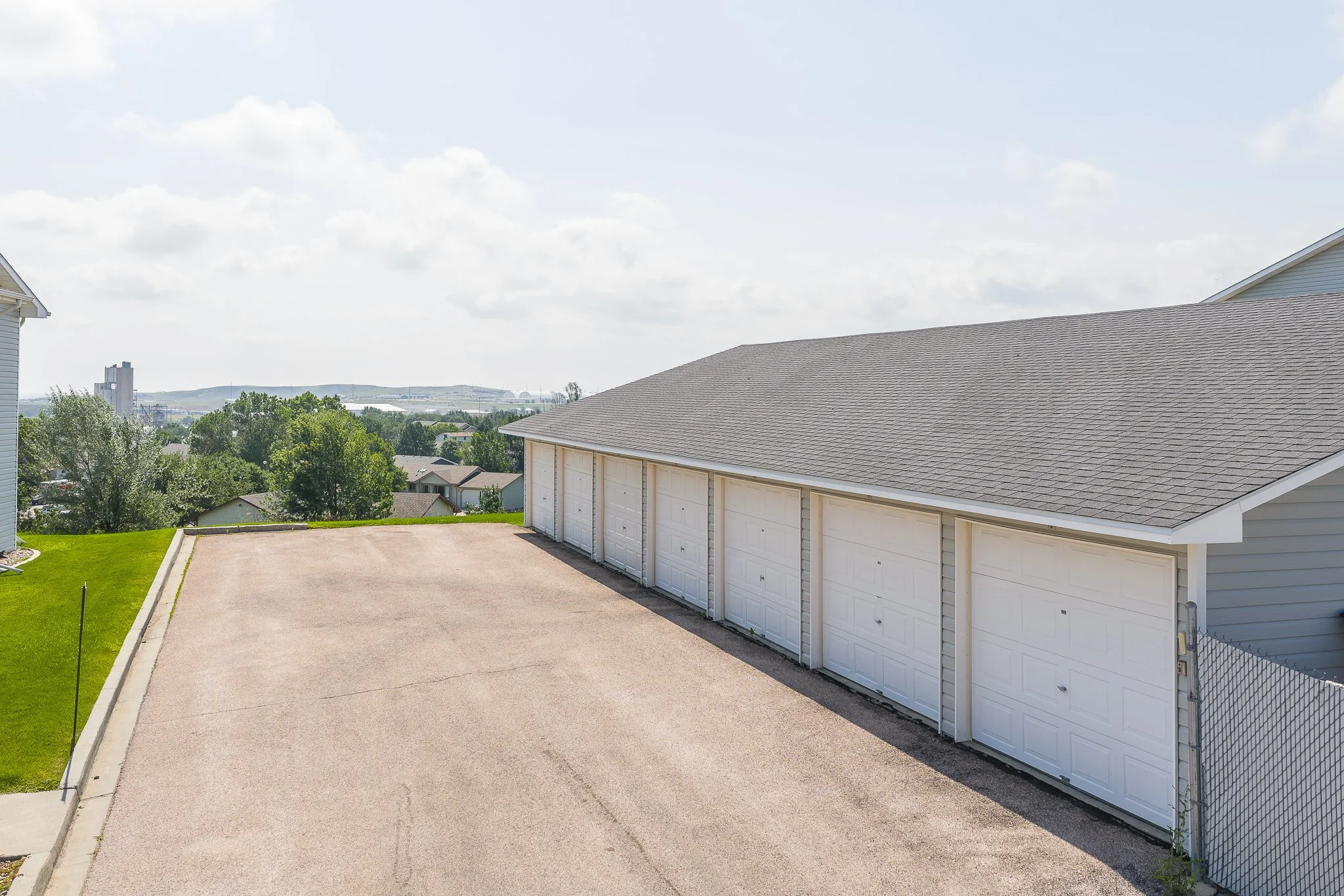 Empty outdoor parking lot next to a building with multiple white garage doors, grassy area, trees, and distant cityscape under partly cloudy sky.