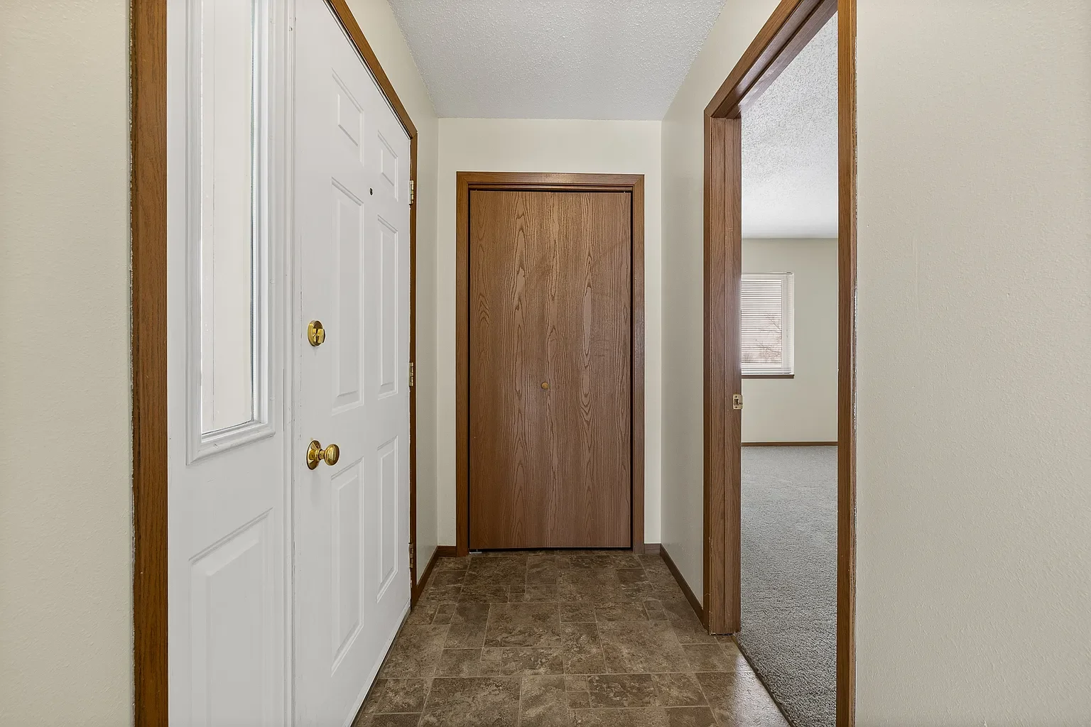 View of an entryway with a closed brown front door, a white closet door with a window, and an open doorway leading into a room with a window and carpeted floor.