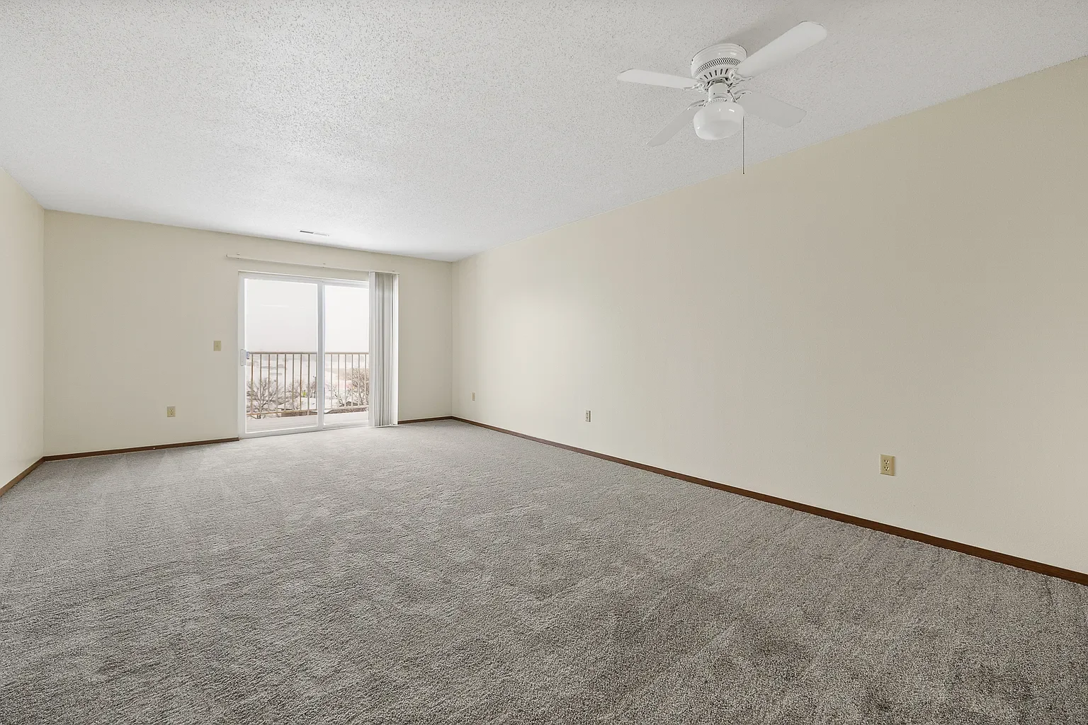 Empty living room with beige walls, gray carpet, sliding glass door to balcony, ceiling fan, and vertical blinds.