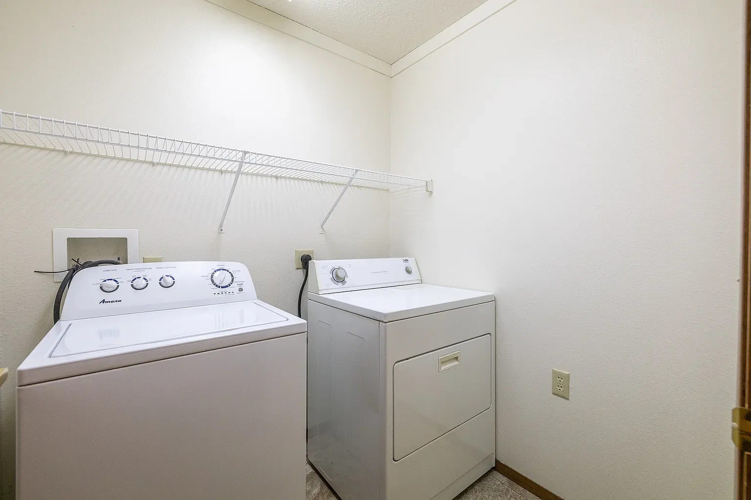 A small laundry room with a white washing machine and a white dryer, a wire shelf above them, a wall outlet, and plain white walls.