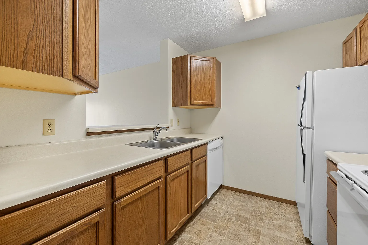 Kitchen with wooden cabinets, a white refrigerator, a double sink, beige countertops, and tiled flooring.