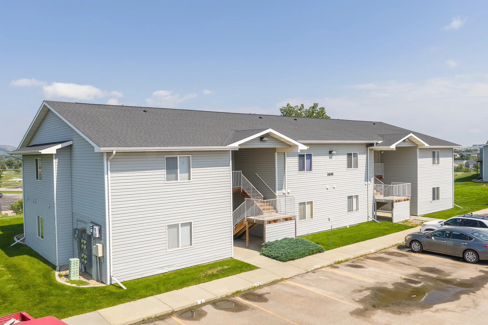 Exterior view of a multi-unit apartment building with white siding, black shingle roof, and small balconies, situated next to a parking lot with cars and patches of water on the ground.